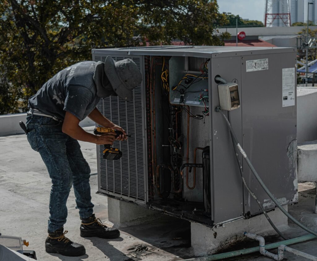 A worker in a bucket hat repairs an outdoor air conditioning unit on a rooftop. HVAC Technician