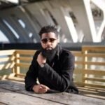 Stylish man sitting on a wooden deck by a bridge, wearing sunglasses and a watch.